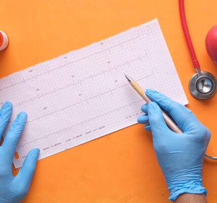 Hands wearing blue gloves holding a pen pointing at an ECG graph with medical equipment nearby.
