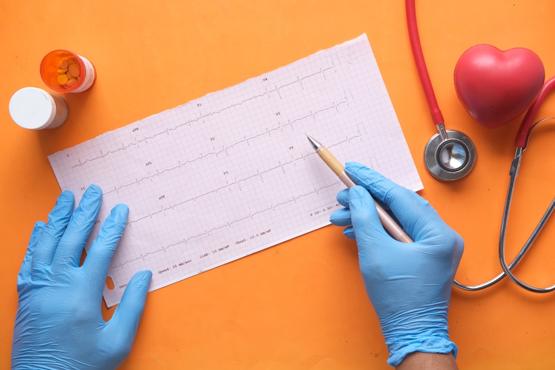 Hands wearing blue gloves holding a pen pointing at an ECG graph with medical equipment nearby.